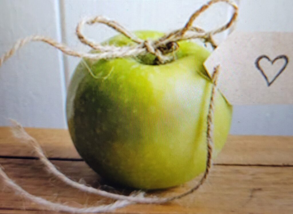 Picture of a Granny smith green apple on a wood table with a string tied in a bow. There is a paper tag attached with an outline of a heart. 