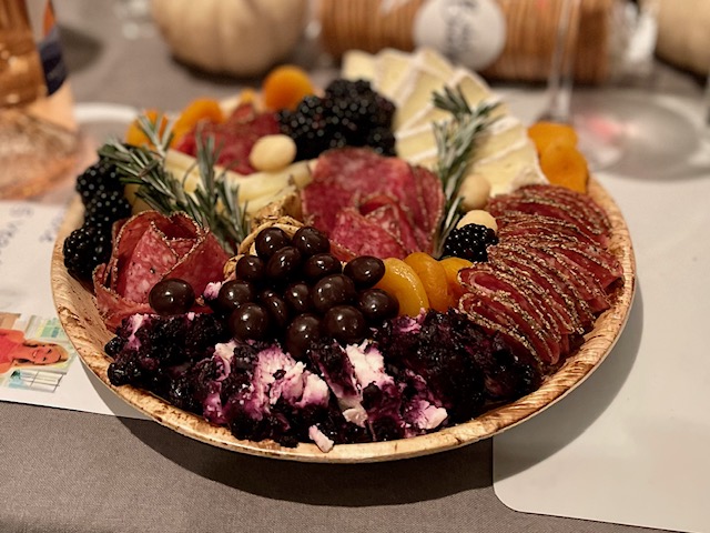 This is a photo of a charcuterie plate.  It is round with herbs, prosciutto, olives, blackberries, cheese (brie, perhaps?) and some kind of orange fruit.  